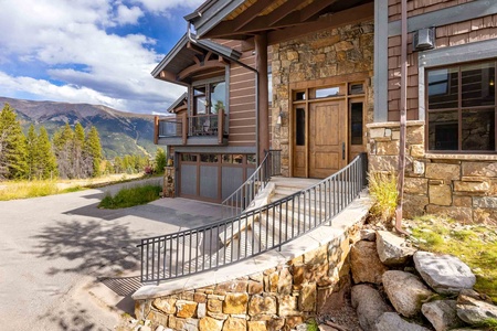 A stone and wood house with a front entrance featuring a wooden door, large windows and garage set against a backdrop of mountains and trees.