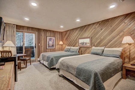 A cozy bedroom with wood-paneled walls, two queen-sized beds, framed artwork, a desk with a chair, and a sliding glass door leading to a balcony.