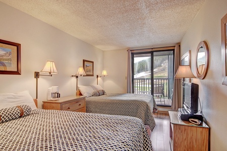 A hotel room with two double beds, bedside lamps, framed pictures, a TV, dresser, and sliding glass door leading to a balcony with an outdoor view.