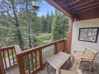 A wooden balcony with chairs and a small table overlooks a forested area with tall pine trees and a creek. There is a decorative wall art piece on the balcony wall.