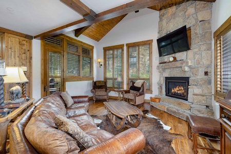 A cozy living room with a leather sectional sofa, stone fireplace, mounted TV, wooden coffee table, and exposed wooden beams on the ceiling. The room has large windows allowing ample natural light in.