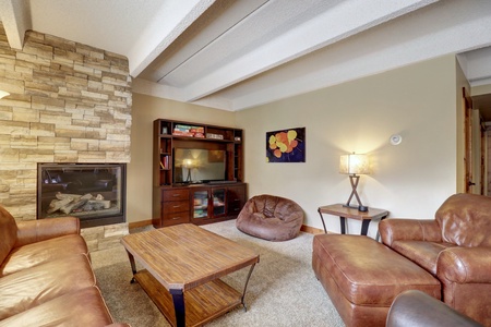 A cozy living room features leather seating, a wooden coffee table, a fireplace with stonework, a TV on a wooden stand, a bean bag chair, and a lamp beside a colorful wall painting.