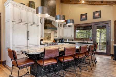 A spacious kitchen features a marble island with eight leather chairs, pendant lights, white cabinetry, stainless steel appliances, and large windows overlooking a wooded area.