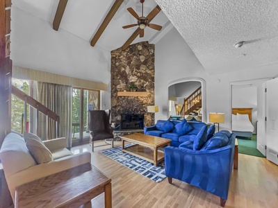 A cozy living room with a high ceiling, wooden beams, and a stone fireplace. It features blue and beige sofas, a wooden coffee table, a ceiling fan, and a view of a lofted bedroom and outdoor greenery.