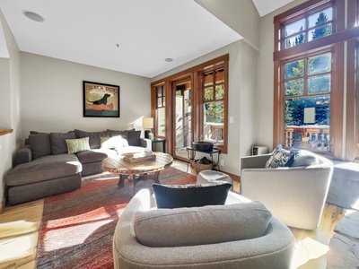 A cozy living room with a sectional sofa, two armchairs, a round coffee table, and large windows offering a view of trees. A framed picture of a dog hangs on the wall above the sofa.