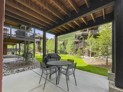 Covered patio area with a round table and four chairs beneath a wooden roof. Nearby are residential buildings and well-maintained grassy areas with trees.