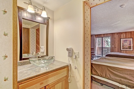 A hotel room with a view of the main bedroom featuring two beds. In the foreground, there is a small vanity area with a glass sink, mirror, and a wall-mounted hairdryer. The room has wooden accents.