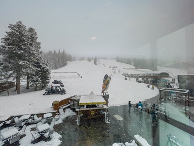 Snow-covered ski resort with a ski slope, trees, and equipment visible. Some people and structures are seen in the foreground with tables and chairs.