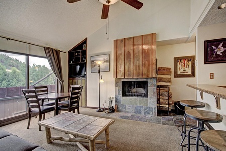 A cozy living room with a fireplace, TV, dining table, sofa, and a wooden coffee table. Large windows with drapes offer a view of trees. A ceiling fan hangs above, and a bar area with stools is on the right.