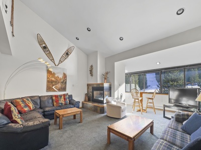 A living room with a blue sectional sofa, wooden coffee table, wall-mounted snowshoes, and a framed painting. There's a fireplace, armchair, TV, and a dining table with chairs by large windows.