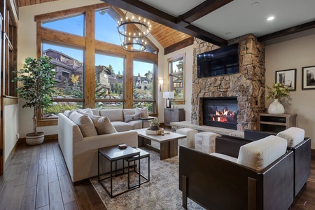 A modern living room with large windows, a stone fireplace, and updated furniture. A round chandelier hangs from the wooden ceiling. A television is mounted above the fireplace.