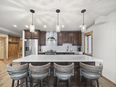 Modern kitchen with a large white island counter, four cushioned bar stools, stainless steel appliances, dark wooden cabinets, and three pendant lights. Hardwood floors and a wooden accent wall are visible.