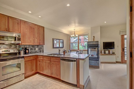 A modern kitchen with wooden cabinets, stainless steel appliances, and a granite countertop opens to a living space with a fireplace, wall-mounted TV, and large window.