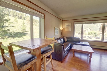 A cozy living room with a black leather sectional sofa, wooden coffee table, and a high dining table with two stools. Large windows offer a scenic view of trees and hills.