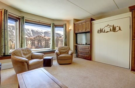 A living room with large windows offering a mountain view, two beige leather chairs, a wooden table, a TV, and a Murphy bed with a mountain and trees design on the front.