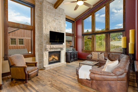 A cozy living room with a stone fireplace, leather furniture, large windows, and wooden floors. A TV is mounted above the fireplace and a ceiling fan hangs from the wooden-beamed ceiling.