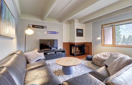 A cozy living room features a leather couch, round wooden coffee table, TV, fireplace, and window with a scenic view. The room is decorated with modern furnishings and a wall-mounted reading lamp.