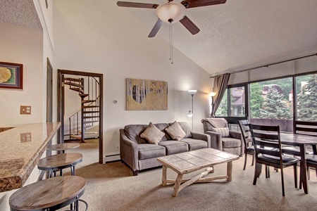 A spacious living room features a gray sofa, a wooden coffee table, a dining table with chairs, and a ceiling fan. A spiral staircase is visible in the background, along with large windows and artwork on the walls.