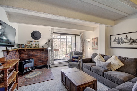 A cozy living room features a brick fireplace, gray sofas with pillows, a wooden coffee table, large sliding glass doors, and wall art. A TV is mounted on the wall.