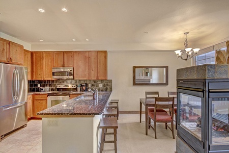 Modern kitchen and dining area with wooden cabinets, stainless steel appliances, granite countertops, an island with bar stools, a dining table with chairs, and a double-sided fireplace.