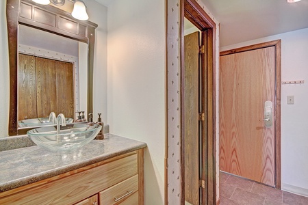 A bathroom with a wooden vanity, glass sink, mirror, and light fixture. The door to the bathroom is open, showing a hallway with closed wooden doors and tiled floor.