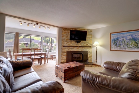 A cozy living room with leather sofas, a wooden trunk coffee table, a fireplace, and a wall-mounted TV. Large windows offer a view of an outdoor dining area with mountains in the background.