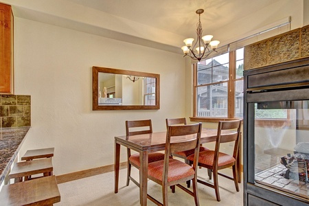 A dining area with a wooden table, four chairs with red cushions, a wall-mounted mirror, a chandelier, and a large window with a view of an outdoor area. Kitchen countertops are partially visible.