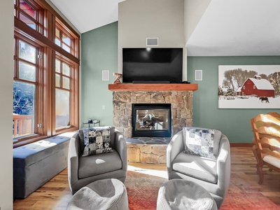 A living room with a stone fireplace, a television above it, two gray armchairs, a painting of a red barn on the wall, and large windows letting in natural light.