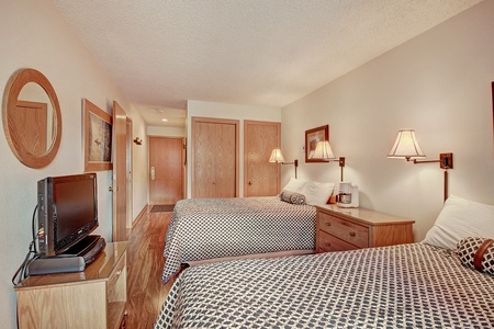 A hotel room with two double beds, a small flat-screen TV on a dresser, wall-mounted lamps, and wooden furnishings. The room has white walls, patterned bed covers, and a wooden floor.