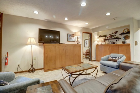 A cozy living room with a mounted TV, a couch, armchairs, a coffee table, shelves with decorative items, a lamp, and a fire extinguisher on the wall. Neutral color palette with soft lighting.