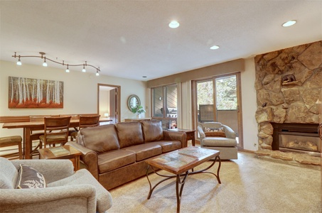 Living room with leather sofa, armchairs, coffee table, stone fireplace, and art on wall. Dining area with wooden table and chairs. Large windows showing an outdoor view.