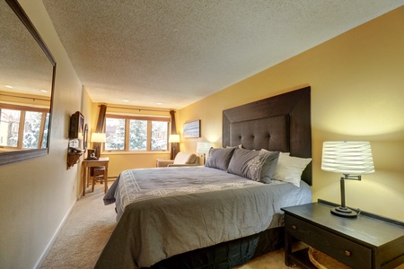 A bedroom with a large bed, gray bedspread, two bedside tables with lamps, a mirror on the left wall, a chair by the window, and a TV mounted on the wall. The walls are painted yellow.