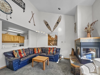 A living room with a blue sectional sofa, colorful throw pillows, and a wooden coffee table. There's a fireplace, wall art, and rustic decor like snowshoes and paddles mounted on the walls.