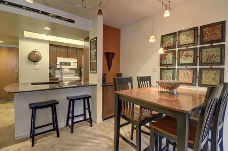 Modern kitchen and dining area with a wooden table and chairs. The kitchen features a white stove and microwave, with a black countertop bar and two stools. Decorative artwork adorns the walls.