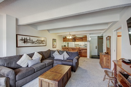 A cozy living room features a gray sectional sofa, wooden coffee table, wall art, and a mounted TV. In the background, there is an open kitchen and dining area with wooden furniture and ceiling beams.