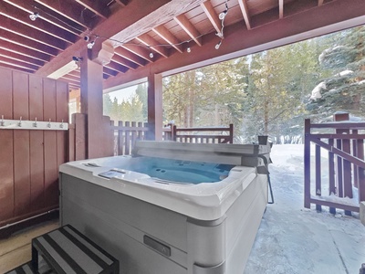 A hot tub under a wooden pergola, surrounded by snow-covered trees. Steam rises from the water, with string lights hanging from the beams above. Steps lead up to the hot tub.