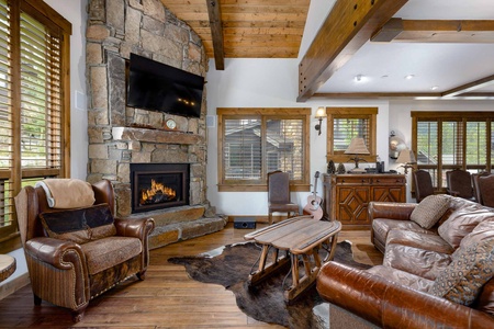 A rustic living room features a stone fireplace with a TV above, leather chairs, a leather sofa, a wooden coffee table on a cowhide rug, and wooden ceiling beams. Large windows offer ample natural light.