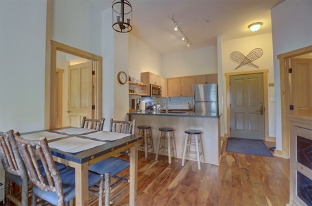 A cozy kitchen with wooden cabinets, stainless steel appliances, and a breakfast bar with stools. Dining table with rustic chairs in foreground, and snowshoes decor on the wall near the entrance door.