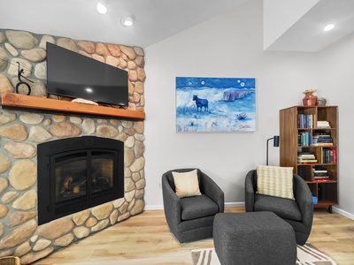 A cozy living room with a stone fireplace, TV mounted above, two grey armchairs, a small ottoman, and a bookshelf. A painting of a blue animal hangs on the wall.