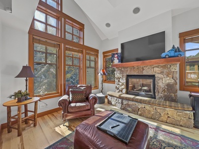 A cozy living room with large windows, a stone fireplace, a leather armchair, and a TV mounted above the mantel. A side table with a lamp and decorative items are also present.