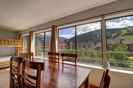 A dining area with a wooden table and chairs is situated near a large window showcasing a scenic mountain view. A bunk bed is visible in the corner to the left.