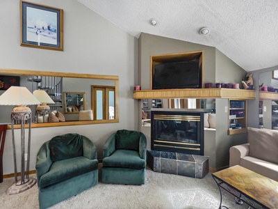 A living room with two green chairs, a beige couch, a fireplace with a TV above it, a large framed mirror, lamps, and wall decor. The carpet is light-colored, and the walls are painted beige.