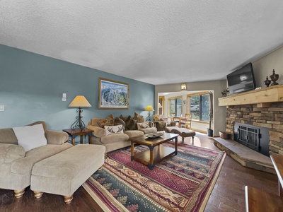 A cozy living room with a couch, armchair, and coffee table on a patterned rug. A stone fireplace sits to the right, and there is a large window and a door leading outside. A painting hangs on the wall.