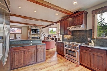 A modern kitchen with wooden cabinets, stainless steel stove, and granite countertops. The room features a beamed ceiling and a view of a living area with a stone fireplace and large windows.