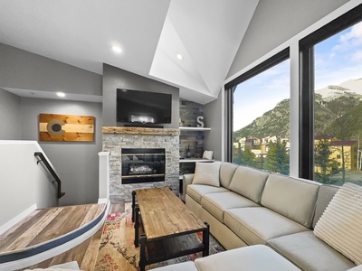 A modern living room with TV mounted above a stone fireplace, large sectional sofa, coffee table, large windows with mountain views.