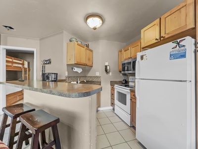 A small kitchen with wooden cabinets, a white refrigerator, a microwave, a stove, and a tiled countertop with two bar stools. Adjacent room visible with bunk beds.