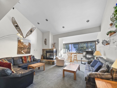 A living room with high ceilings, a fireplace, various seating, a wooden coffee table, a television, and wall-mounted decorations including snowshoes and a landscape painting. Large window at the back.