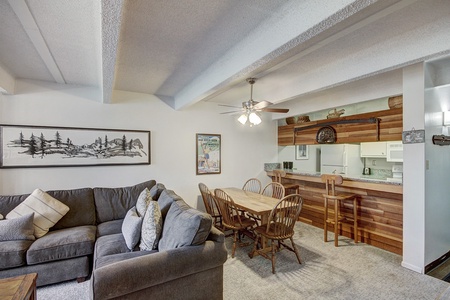 A living room with a grey sectional sofa, pillows, and wall art, adjacent to a dining area featuring a wooden table with six chairs and a kitchen with wooden cabinets and a ceiling fan.