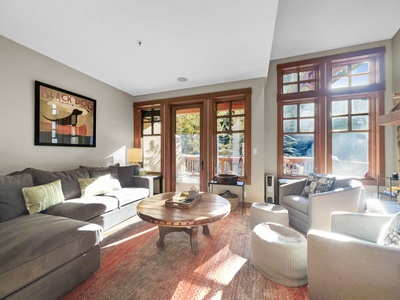 A cozy living room features a sectional sofa, two armchairs, a round wooden coffee table, large windows with wooden frames, and a framed dog poster on the wall.