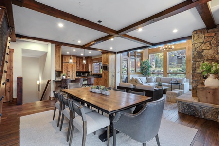 A modern living and dining area with wooden beams, a large dining table, comfortable seating, a stone accent wall, and a view of the outside.